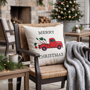 A square throw pillow with a Christmas design sits on a woven outdoor chair. The pillow is cream-colored and features a red vintage truck carrying a Christmas tree with the words 'MERRY CHRISTMAS' printed in black.
