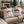 Two cream throw pillows on a beige outdoor sofa, printed with a red vintage truck carrying Christmas trees and the words 'MERRY CHRISTMAS'. The background shows a cozy patio setting with a stone fireplace, green garlands, and potted trees.