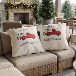 Two cream throw pillows on a beige outdoor sofa, printed with a red vintage truck carrying Christmas trees and the words 'MERRY CHRISTMAS'. The background shows a cozy patio setting with a stone fireplace, green garlands, and potted trees.