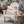 A distressed white wooden Adirondack chair in a snowy outdoor setting, styled with a cream throw pillow featuring a vintage red and blue 'Air Mail' Santa Claus design. A rustic tree stump side table holds wrapped gifts and a copper mug next to the chair.