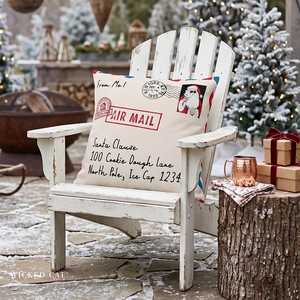 A distressed white wooden Adirondack chair in a snowy outdoor setting, styled with a cream throw pillow featuring a vintage red and blue 'Air Mail' Santa Claus design. A rustic tree stump side table holds wrapped gifts and a copper mug next to the chair.