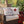 Two decorative throw pillows styled on a wicker outdoor sofa, designed to look like vintage airmail and envelope mailers with red, white, and blue details. A red plaid throw blanket is draped over the arm, and a decorated Christmas tree is visible in the background.