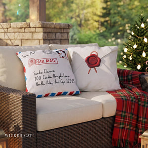 Two decorative throw pillows styled on a wicker outdoor sofa, designed to look like vintage airmail and envelope mailers with red, white, and blue details. A red plaid throw blanket is draped over the arm, and a decorated Christmas tree is visible in the background.