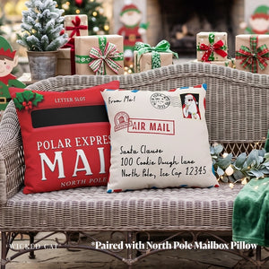 Two Christmas-themed decorative throw pillows displayed on a wicker sofa in a holiday setting. One pillow is red with a black letter slot graphic and white 'POLAR EXPRESS MAIL' text, and the other is cream with an 'AIR MAIL' stamp design, Santa portrait, and address text.
