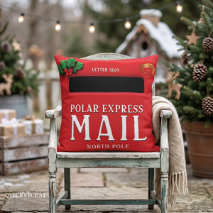 A red decorative throw pillow styled on a distressed white wooden chair in a cozy, festive outdoor setting. The pillow features a 'Polar Express Mail' design with a black letter slot graphic, white lettering, a holly leaf accent, and a small gold seal.
