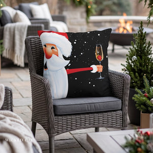 A square decorative throw pillow featuring a festive Christmas design of Santa Claus holding a champagne glass, resting on a gray wicker outdoor patio chair.