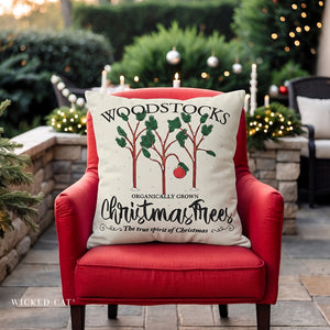 A square throw pillow with a Christmas tree graphic and the text 'WOODSTOCKS Organically Grown Christmas Trees' resting on a red armchair in a decorated outdoor patio setting.