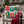 A square holiday throw pillow with a vintage postage stamp collage design featuring Christmas drinks and messages, resting on a rustic wooden chair with a red knit blanket draped over the arm in an outdoor patio setting.