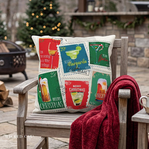 A square holiday throw pillow with a vintage postage stamp collage design featuring Christmas drinks and messages, resting on a rustic wooden chair with a red knit blanket draped over the arm in an outdoor patio setting.