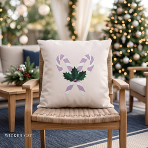 A square cream throw pillow with a Christmas design of two purple-and-white candy canes crossing over green holly leaves, displayed on a woven wooden chair in a cozy, softly blurred living room with a Christmas tree in the background.