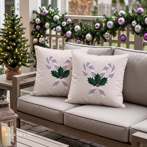 Two beige outdoor throw pillows with a green holly and purple candy cane design, displayed on a beige patio sofa decorated for Christmas.