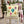 A square decorative throw pillow with a pastel yellow background and colorful hanging Christmas ornament illustrations, displayed on a distressed white wooden chair with a plaid blanket in a holiday outdoor setting.