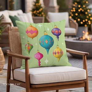 A square decorative throw pillow with a pastel yellow background and colorful hanging Christmas ornament illustrations, displayed on a distressed white wooden chair with a plaid blanket in a holiday outdoor setting.