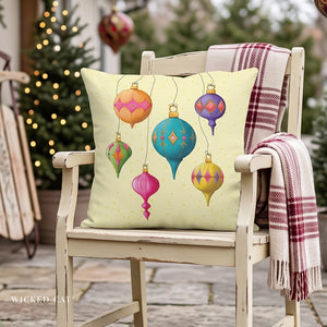 A square decorative throw pillow with a pastel yellow background and colorful hanging Christmas ornament illustrations, displayed on a distressed white wooden chair with a plaid blanket in a holiday outdoor setting.