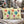 Two square decorative throw pillows with a festive Christmas ornament print, displayed on a gray outdoor patio chair with a knitted blanket, set against a blurred background of decorated Christmas trees and a fire pit.