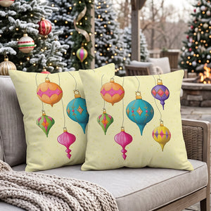 Two square decorative throw pillows with a festive Christmas ornament print, displayed on a gray outdoor patio chair with a knitted blanket, set against a blurred background of decorated Christmas trees and a fire pit.