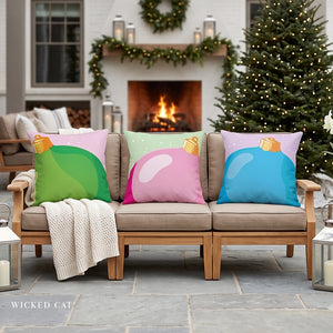 Three decorative outdoor throw pillows with colorful Christmas ornament designs are arranged on a beige patio sofa in front of a lit fireplace. The pillows feature green, pink, and blue ornaments, set against a cozy holiday background with a Christmas tree and garlands.