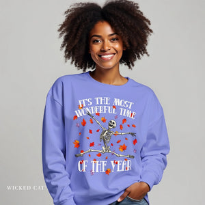 A smiling woman with curly dark hair wears a lavender crewneck sweatshirt featuring a skeleton dancing among red and orange autumn leaves with the text 'IT'S THE MOST WONDERFUL TIME OF THE YEAR'. The brand name 'Wicked Cat' is visible in the bottom-left corner.