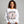 A smiling woman wearing a white crewneck sweatshirt with a Halloween graphic of a skeleton on a broomstick surrounded by falling autumn leaves. The text reads 'IT'S THE MOST WONDERFUL TIME OF THE YEAR'.