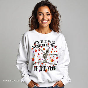 A smiling woman wearing a white crewneck sweatshirt with a Halloween graphic of a skeleton on a broomstick surrounded by falling autumn leaves. The text reads 'IT'S THE MOST WONDERFUL TIME OF THE YEAR'.