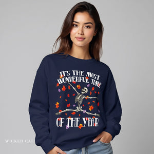 A woman wearing a navy blue crewneck sweatshirt with a graphic of a skeleton sitting cross-legged surrounded by colorful autumn leaves, with the text 'IT'S THE MOST WONDERFUL TIME OF THE YEAR'.