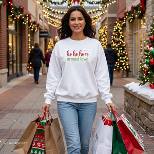 Woman carrying shopping bags walking down a Christmas street wearing a white sweatshirt that reads ho ho ho'n around town in red and green text.