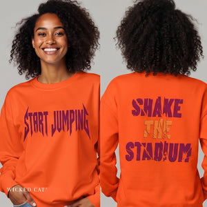 An orange crewneck sweatshirt shown on a model from both the front and back views. The front features the text 'START JUMPING' in a dark purple stylized font, while the back displays 'SHAKE THE STADIUM?' in a distressed purple font with a small gold accent in the middle word.