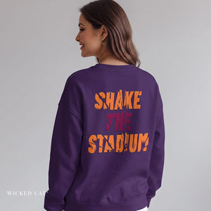 A woman shown from the back wearing a purple crewneck sweatshirt with 'SHAKE THE STADIUM' printed in orange and red across the back. The brand name 'WICKED CAT' is visible in the bottom-left corner.