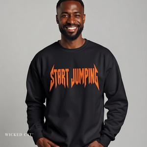 A smiling man wearing a black crewneck sweatshirt with a bold orange graphic text reading 'START JUMPING' across the chest, set against a plain gray studio background.