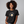 A woman with curly brown hair wearing a black graphic t-shirt that reads 'THIS IS NOT AN EGG' around a frying pan illustration with a yellow egg yolk. The brand name 'Wicked Cat' is visible in the bottom-left corner against a solid gray studio background.