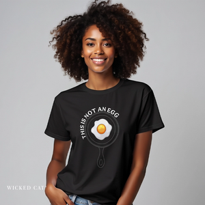 A woman with curly brown hair wearing a black graphic t-shirt that reads 'THIS IS NOT AN EGG' around a frying pan illustration with a yellow egg yolk. The brand name 'Wicked Cat' is visible in the bottom-left corner against a solid gray studio background.
