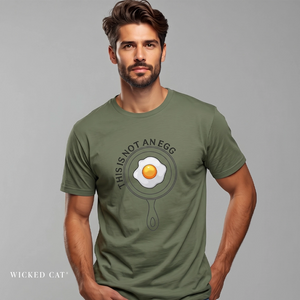 A man wearing an olive green t-shirt with a graphic of a frying pan containing an egg, with the text 'THIS IS NOT AN EGG' printed around it. The brand name 'WICKED CAT' is visible in the bottom-left corner.