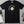 A black short-sleeve graphic T-shirt laid flat on a gray surface, featuring a frying pan illustration with a fried egg and the text 'THIS IS NOT AN EGG.' Styled flat-lay props around it include a mug, spoon, book, glasses, and a wallet.