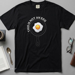 A black short-sleeve graphic T-shirt laid flat on a gray surface, featuring a frying pan illustration with a fried egg and the text 'THIS IS NOT AN EGG.' Styled flat-lay props around it include a mug, spoon, book, glasses, and a wallet.