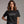 A woman wearing a black short-sleeve graphic T-shirt with white text that reads, 'Rain is just God crying, probably because of something you did.' The shirt is shown against a plain gray studio background, with the brand name 'WICKED CAT' visible in the lower left corner.