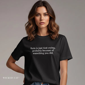 A woman wearing a black short-sleeve graphic T-shirt with white text that reads, 'Rain is just God crying, probably because of something you did.' The shirt is shown against a plain gray studio background, with the brand name 'WICKED CAT' visible in the lower left corner.