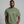 A man wearing an olive green t-shirt with white printed text that reads 'Rain is just God crying, probably because of something you did.' against a solid gray background.