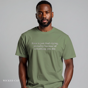 A man wearing an olive green t-shirt with white printed text that reads 'Rain is just God crying, probably because of something you did.' against a solid gray background.