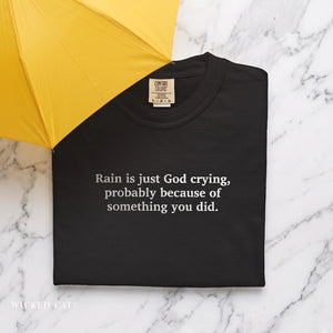 A folded black t-shirt with white text reading 'Rain is just God crying, probably because of something you did.' displayed on a white marble background with a yellow umbrella in the top-left corner.