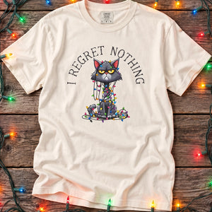 t-shirt with a cartoon cat tangled in Christmas lights with text at the top that says I Regret Nothing, laying on a wood floor with christmas lights around it.