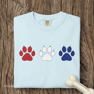 Light blue t-shirt with red, white, and blue paw prints on a wooden surface.