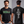 A man modeling a black graphic T-shirt shown from both front and back. The front has a green distressed street-sign style graphic reading 'ELM STREET.