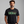 A man wearing a black crewneck t-shirt with a distressed green 'ELM STREET' sign graphic on the chest, set against a plain gray studio background.