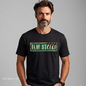 A man wearing a black crewneck t-shirt with a distressed green 'ELM STREET' sign graphic on the chest, set against a plain gray studio background.