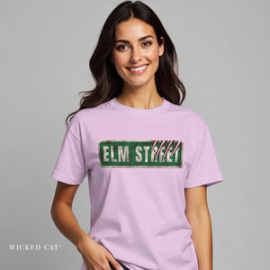 An lilac short-sleeve crewneck t-shirt featuring a distressed green 'ELM STREET' sign graphic printed across the chest.
