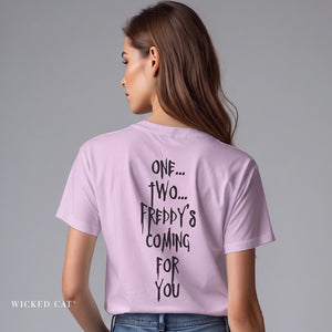 A woman shown from the back wearing a light lavender-purple t-shirt with black hand-drawn text on the back reading 'ONE TWO FREDDYS COMING FOR YOU