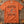 An orange crewneck t-shirt featuring a Halloween graphic of a dancing skeleton surrounded by falling autumn leaves, with the text 'IT'S THE MOST WONDERFUL TIME OF THE YEAR'. The shirt is displayed flat on a rustic wooden background with scattered leaves.