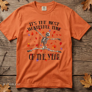 An orange crewneck t-shirt featuring a Halloween graphic of a dancing skeleton surrounded by falling autumn leaves, with the text 'IT'S THE MOST WONDERFUL TIME OF THE YEAR'. The shirt is displayed flat on a rustic wooden background with scattered leaves.