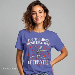 A woman wearing a periwinkle blue graphic t-shirt with a skeleton dancing amidst red and orange autumn leaves, with the text 'IT'S THE MOST WONDERFUL TIME OF THE YEAR'. The brand name 'Wicked Cat' is visible in the bottom-left corner.