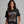 A woman wearing a black graphic t-shirt with a skeleton and autumn leaves design, posed against a solid gray studio background.
